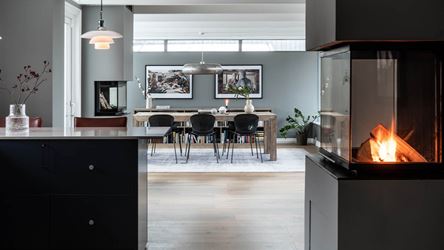 Bildet viser en lekker leilighet med stilfullt interiør. Det er et kjøkkenet med svarte fronter i forkant, en peis lyser opp til høyre i bildet og man ser ut mot spisestuen i bakgrunnen.
