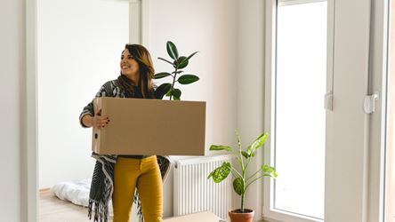 Illustrasjonsbilde av en dame som skal flytte inn i ny leilighet. Hun bærer inn en flyttekasse med en grønn plante som stikker opp av flyttekassen.