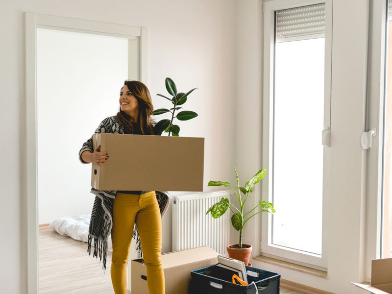 Illustrasjonsbilde av en dame som skal flytte inn i ny leilighet. Hun bærer inn en flyttekasse med en grønn plante som stikker opp av flyttekassen.