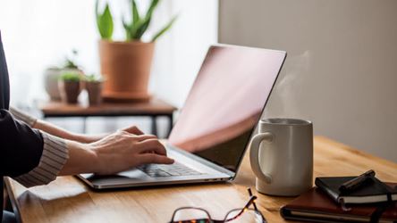 Illustrasjonsbilde av et typisk hjemmekontor. En arbeidsbenk med en laptop, briller og en kopp kaffe. Man ser hendene til en dame som trykker på tastaturet på PCen.