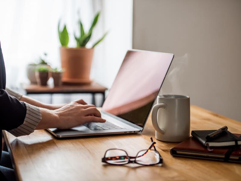 Illustrasjonsbilde av et typisk hjemmekontor. En arbeidsbenk med en laptop, briller og en kopp kaffe. Man ser hendene til en dame som trykker på tastaturet på PCen.