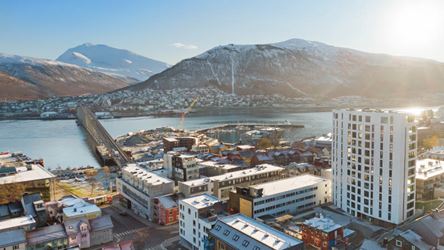 Bildet viser et dronebilde tatt av boligprosjektet Saras Hage og Solseilet i Tromsø. Man ser vakre Tromsø i bakgrunnen med bebyggelse, fjell, vann og blå himmel med strålende sol.