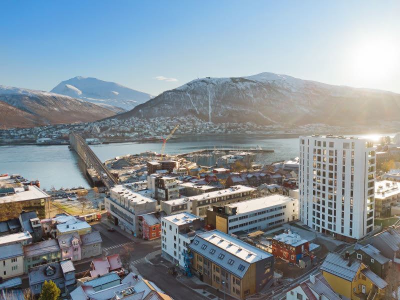 Bildet viser et dronebilde tatt av boligprosjektet Saras Hage og Solseilet i Tromsø. Man ser vakre Tromsø i bakgrunnen med bebyggelse, fjell, vann og blå himmel med strålende sol.
