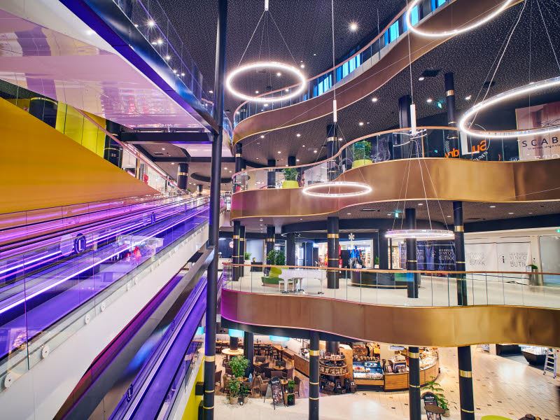 Bilde fra Lagunen Storsenter som viser rulletrapp med lilla lys, store flotte lamper og gulldetaljer i de ulike etasjene i kjøpesenteret.