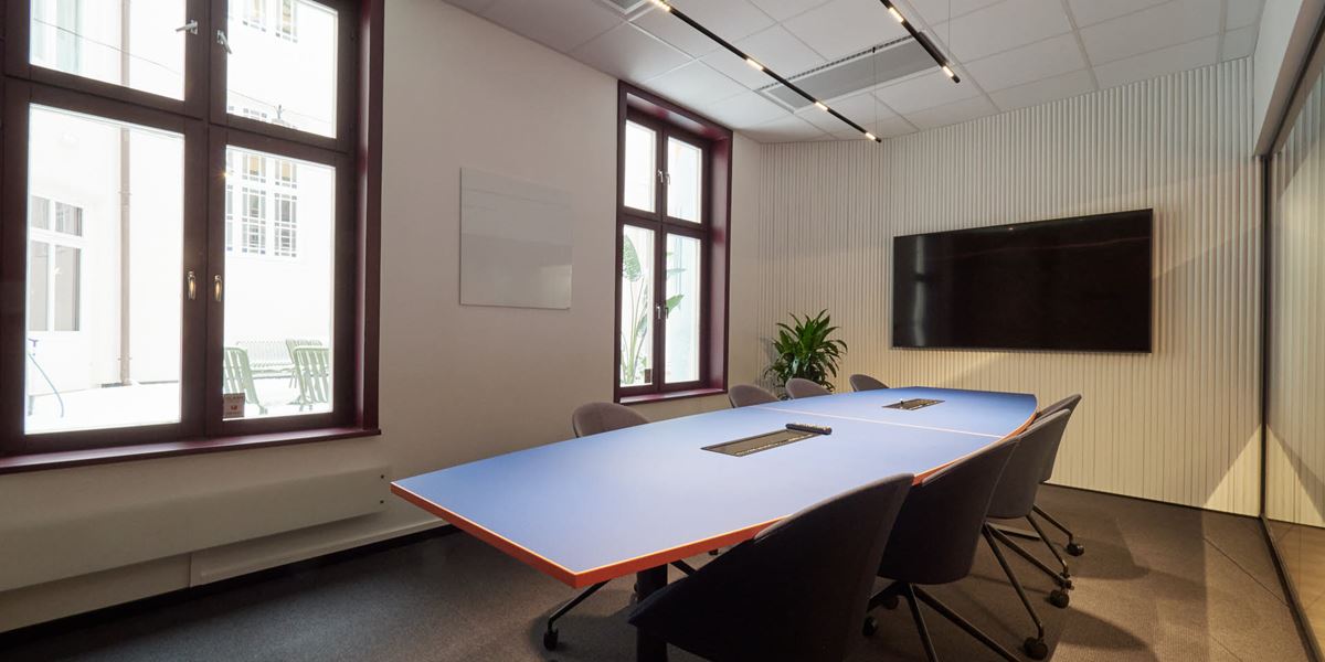 Bilde av et av møterommene i lokalene til Thon Flex i Karl Johans gate 5. Stort møterom med hvite vegger. blått bord,  sorte stoler og en stor TV på veggen.