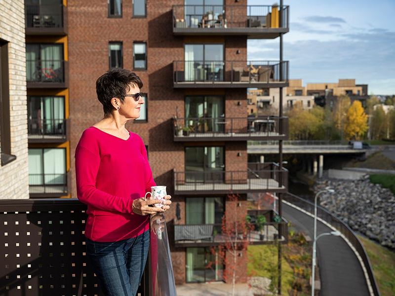 Bildet viser en dame i rød genser som står på balkongen sin i boligprosjektet Strømmen Verksted.