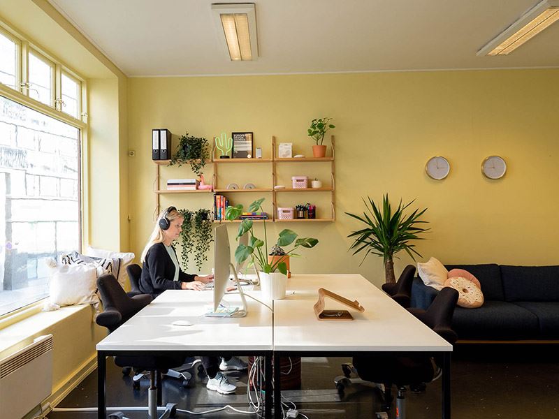 Bildet viser et kontorlokale med gule, kule vegger. Stor hvit kontorpult hvor det sitter en dame som jobber på laptop.